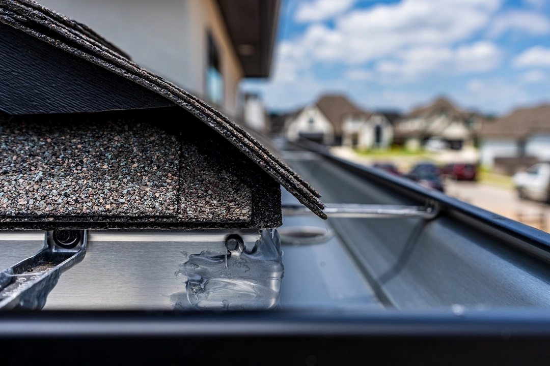 Closeup of asphalt roof shingles and a newly installed metal gutter, with a residential neighborhood blurred in the background.